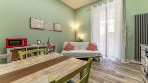 Une pièce comprenant une table à manger, des chaises, un canapé avec des coussins rouges, un four à micro-ondes rouge, des œuvres d’art murales, une lampe et une fenêtre avec des rideaux.