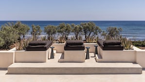 A seaside lounge with three cushioned seating areas, each with a pillow, surrounded by plants and overlooking the ocean. - Exomitis1 Cellar & Sea Escape (Santorini)