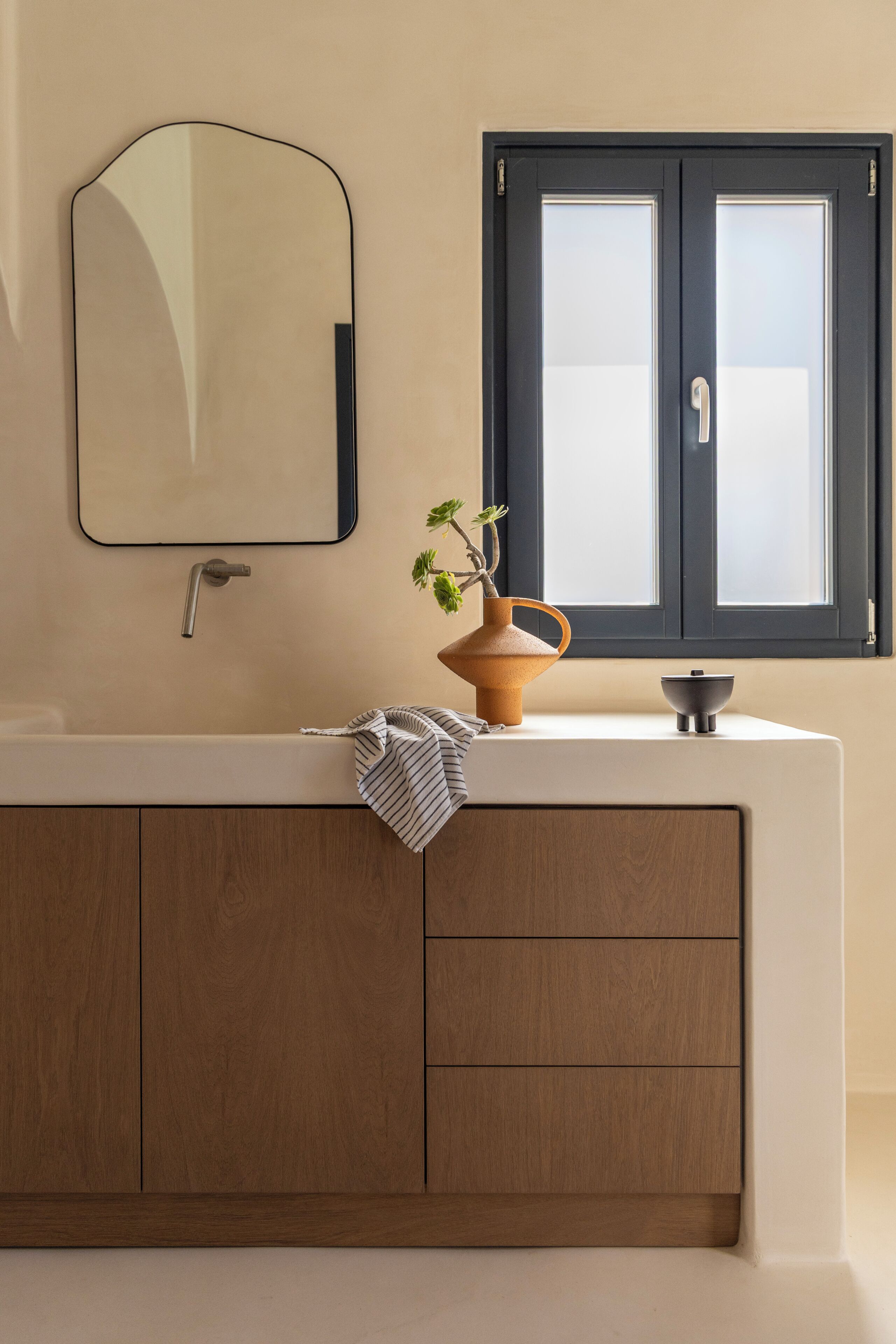 Une salle de bain avec un meuble-lavabo en bois, un miroir, une fenêtre et une plante.
