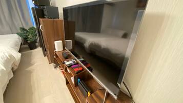 Ein Hotelzimmer mit einem großen Flachbildfernseher, einem Holznachttisch mit Fernbedienung, einem kleinen Lautsprecher und einer Topfpflanze.