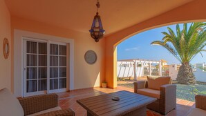 Un patio al aire libre con muebles de mimbre, una mesa, un farol colgante y vistas a edificios y palmeras.