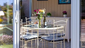 Een glazen terras met een ronde tafel, witte stoelen en een bloevas.
