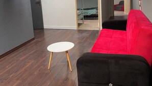 Een moderne kamer met een rode bank, een klein wit tafeltje, een aan de muur bevestigde televisie en een houten deur naar een andere kamer.
