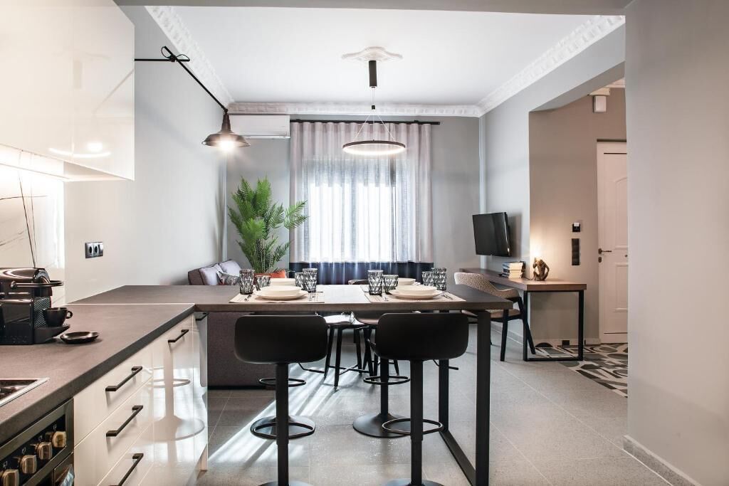 A modern kitchen with a central island, bar stools, and a dining area with a TV and plant.