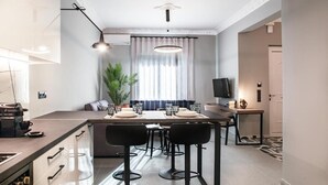 Una cocina moderna con una isla central, taburetes de bar y un comedor con televisor y planta.