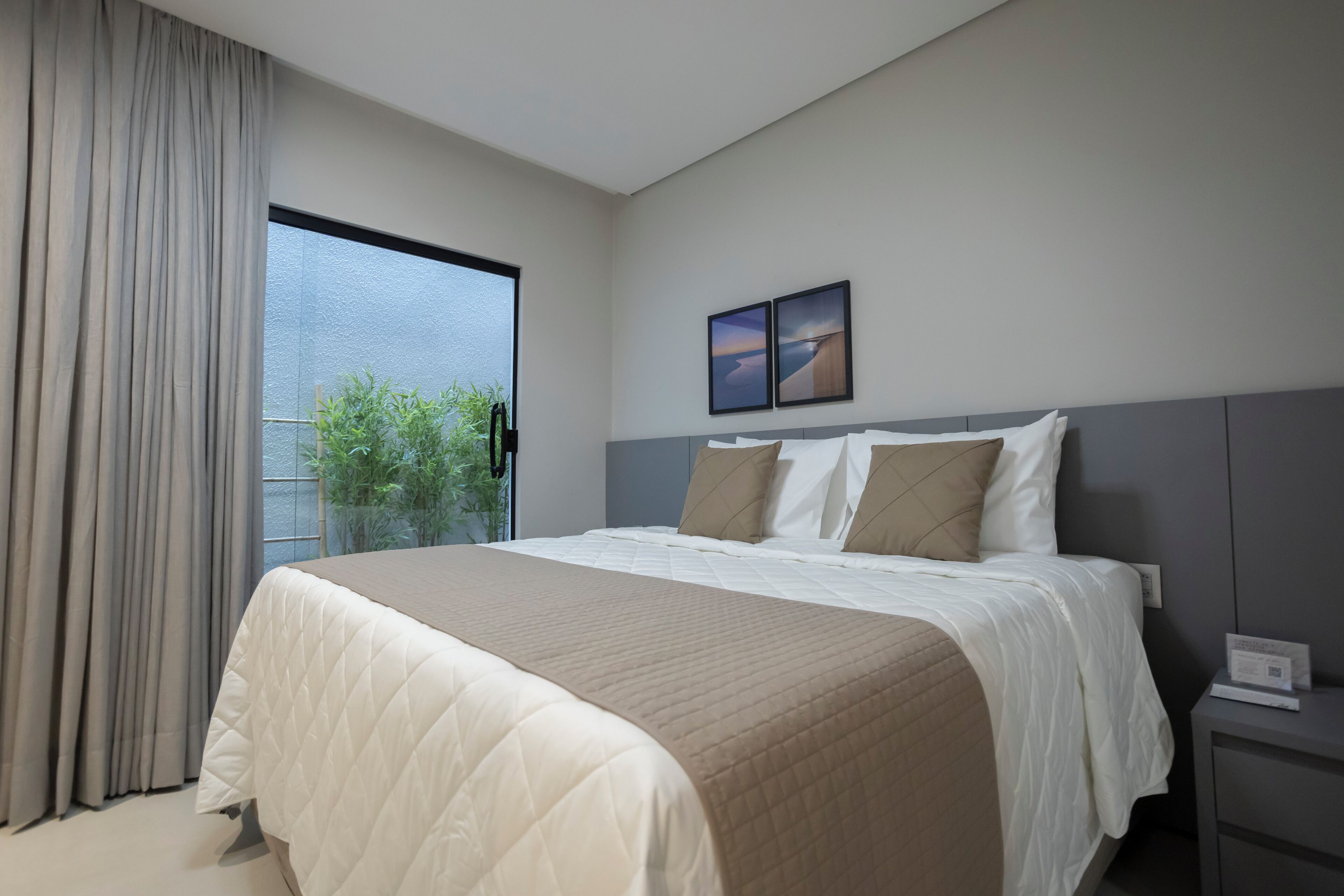 Una habitación de hotel moderna con una cama grande, una mesita de noche, un ventanal con cortinas y dos cuadros en la pared.