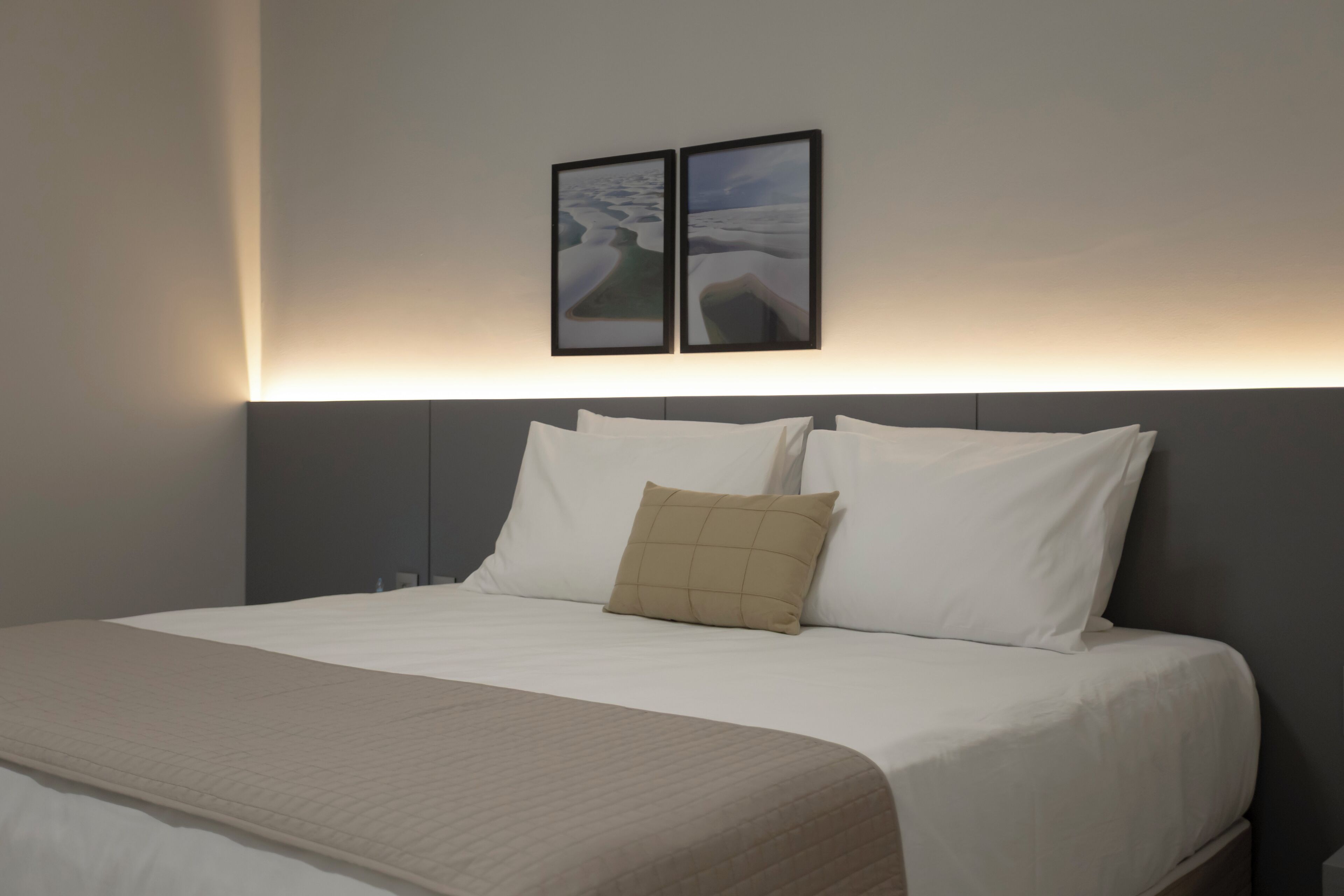 A neatly made bed with white linens and a beige throw blanket, a grey headboard, and two framed pictures on the wall.