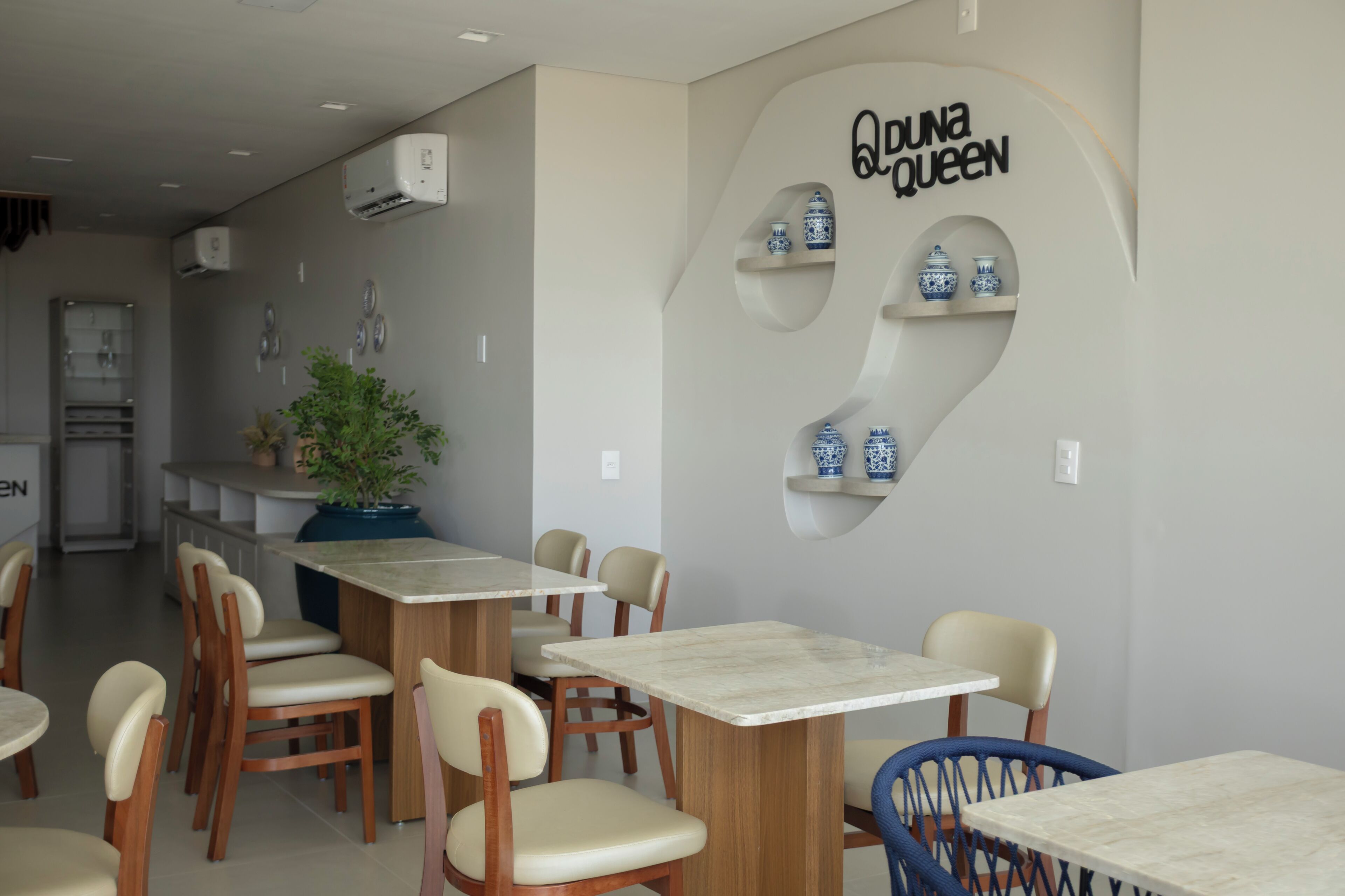 A restaurant interior with wooden tables and chairs, a wall-mounted shelf with blue and white vases, and an air conditioning unit.