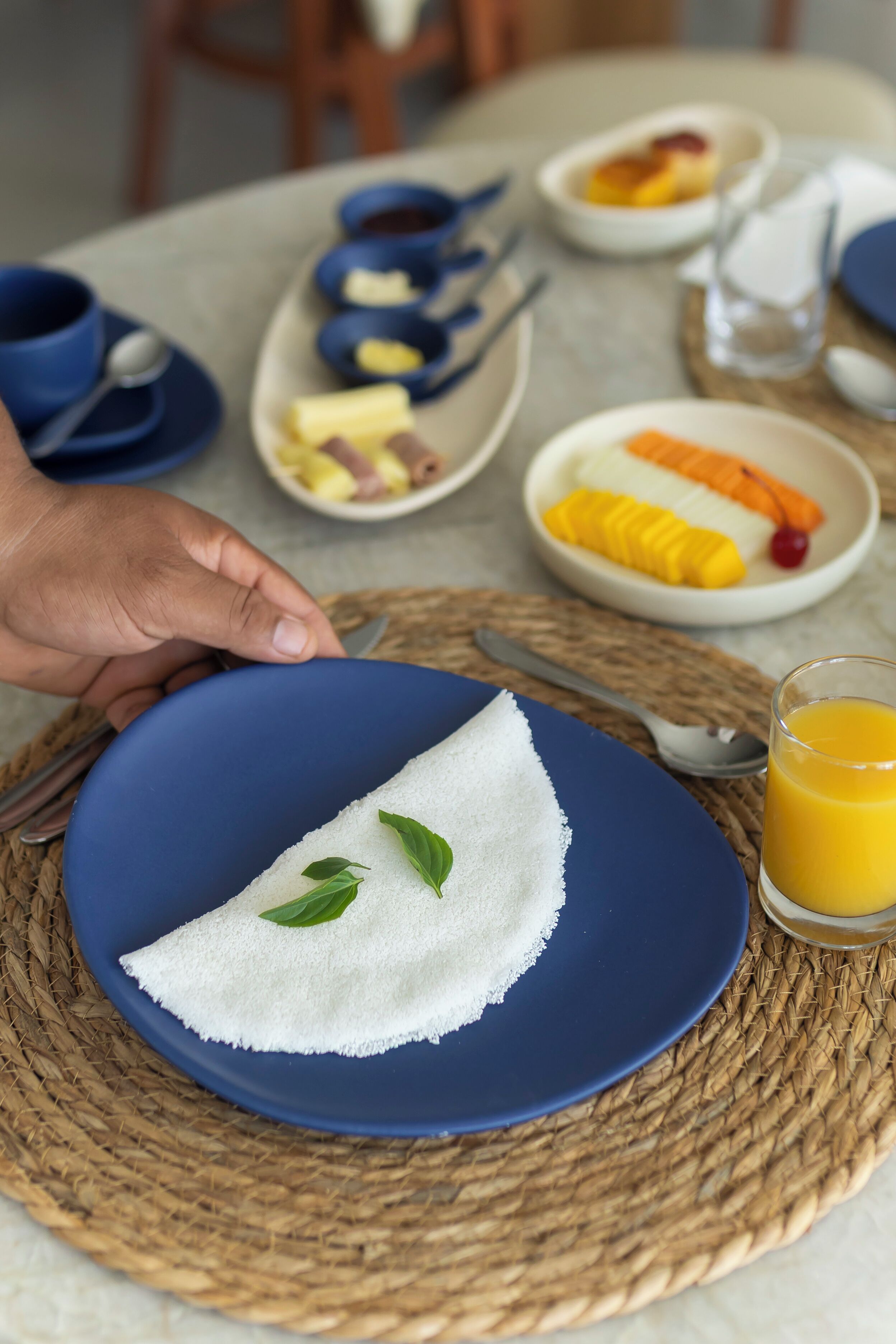 Une table mise pour le petit-déjeuner, avec une assiette bleue, un set de table en fibres tressées et un verre de jus d’orange.