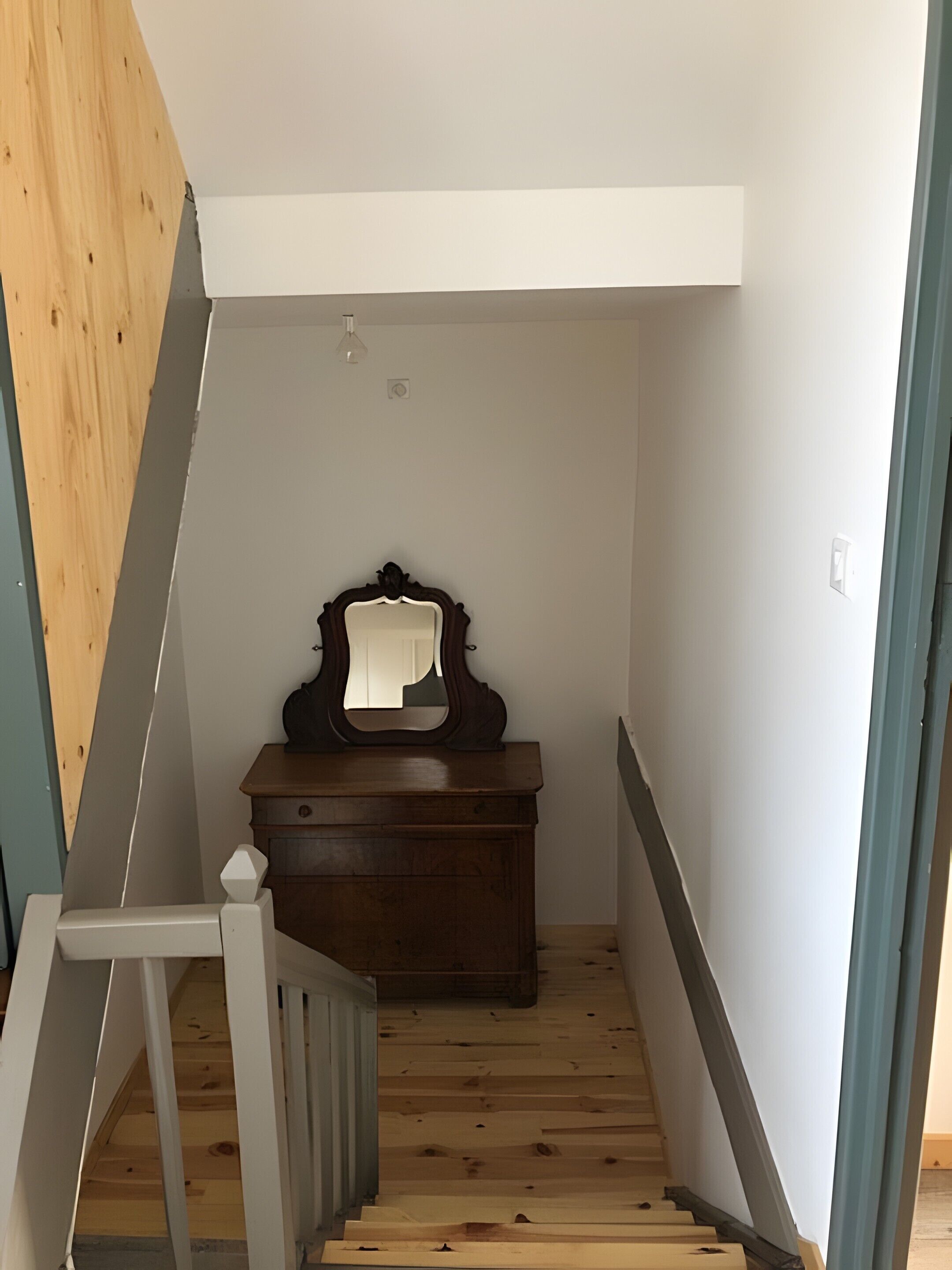 Un escalier étroit avec des marches en bois et une rampe blanche, menant à une pièce où se trouvent une commode en bois et un miroir.