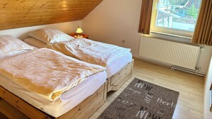 Quarto com teto inclinado, cama com roupa de cama branca, mesa de cabeceira com abajur, janela com cortinas e radiador.