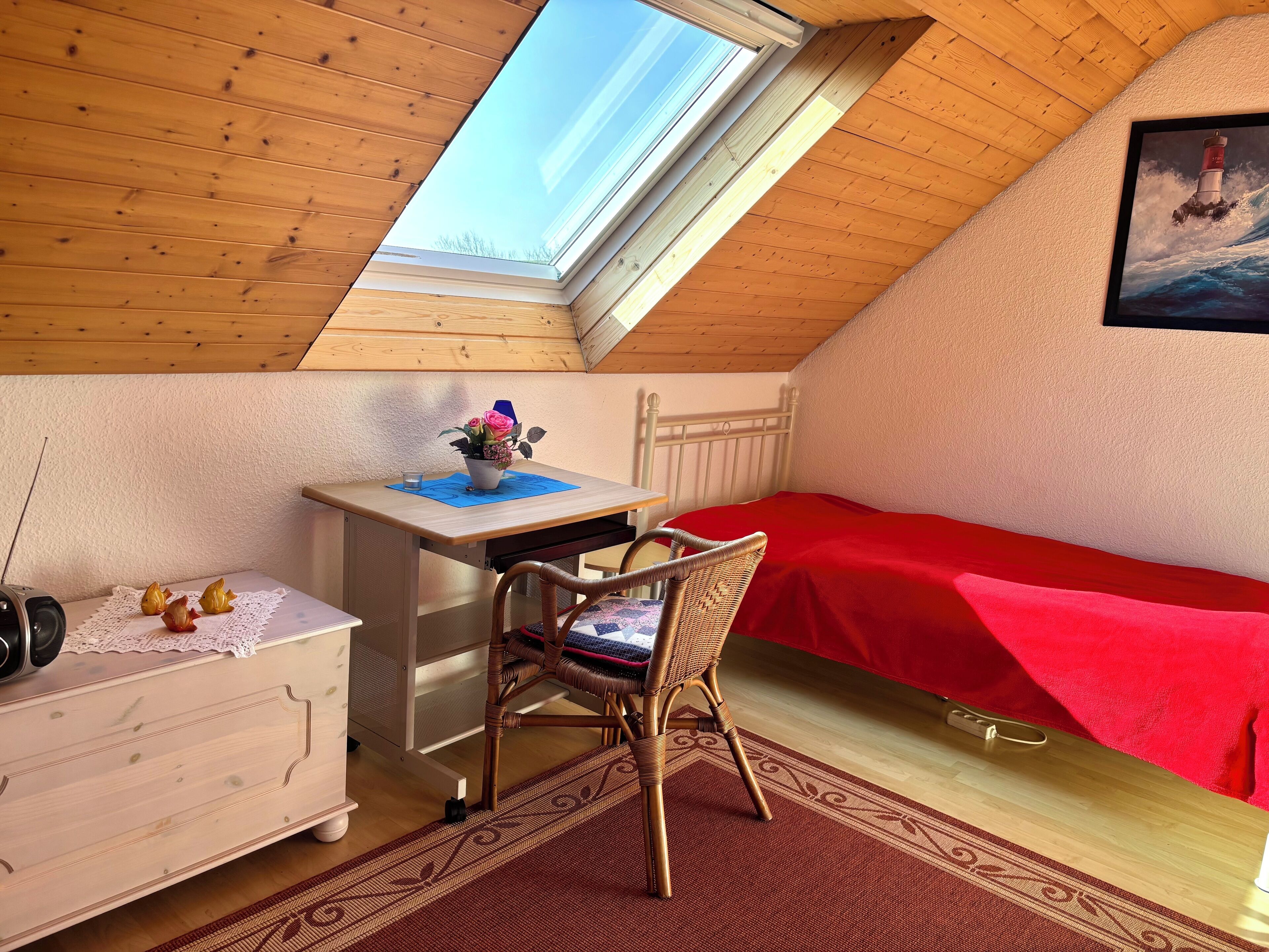 Ein Zimmer mit schrägen Wänden, einem Oberlicht, einem Schreibtisch mit Stuhl, einem Bett mit roter Bettdecke und einem gerahmten Bild an der Wand.