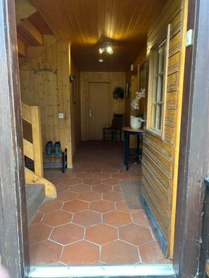 Un couloir intérieur aux murs en bois, doté d’un sol carrelé, avec une porte au bout et une petite table sur laquelle est posée une plante en pot.