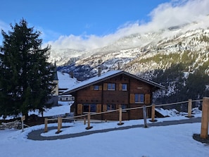 A wooden chalet with a sloped roof, surrounded by snow-covered mountains and a clear sky. - Chalet Mallum – Upper Apartment (Grengiols)