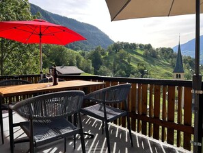 Une terrasse en bois avec une table et des chaises, un parasol rouge et une vue pittoresque sur des collines verdoyantes et le clocher d’une église.