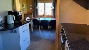 Eine kleine Küche mit Kaffeemaschine, ein Essbereich mit Stühlen und Tisch sowie ein Fenster mit Vorhängen.