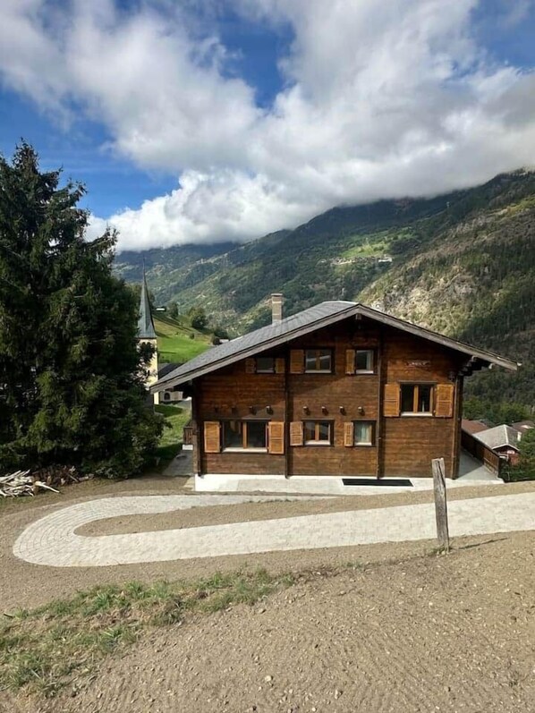 A wooden chalet with a steep roof, surrounded by a mountainous landscape and a church.