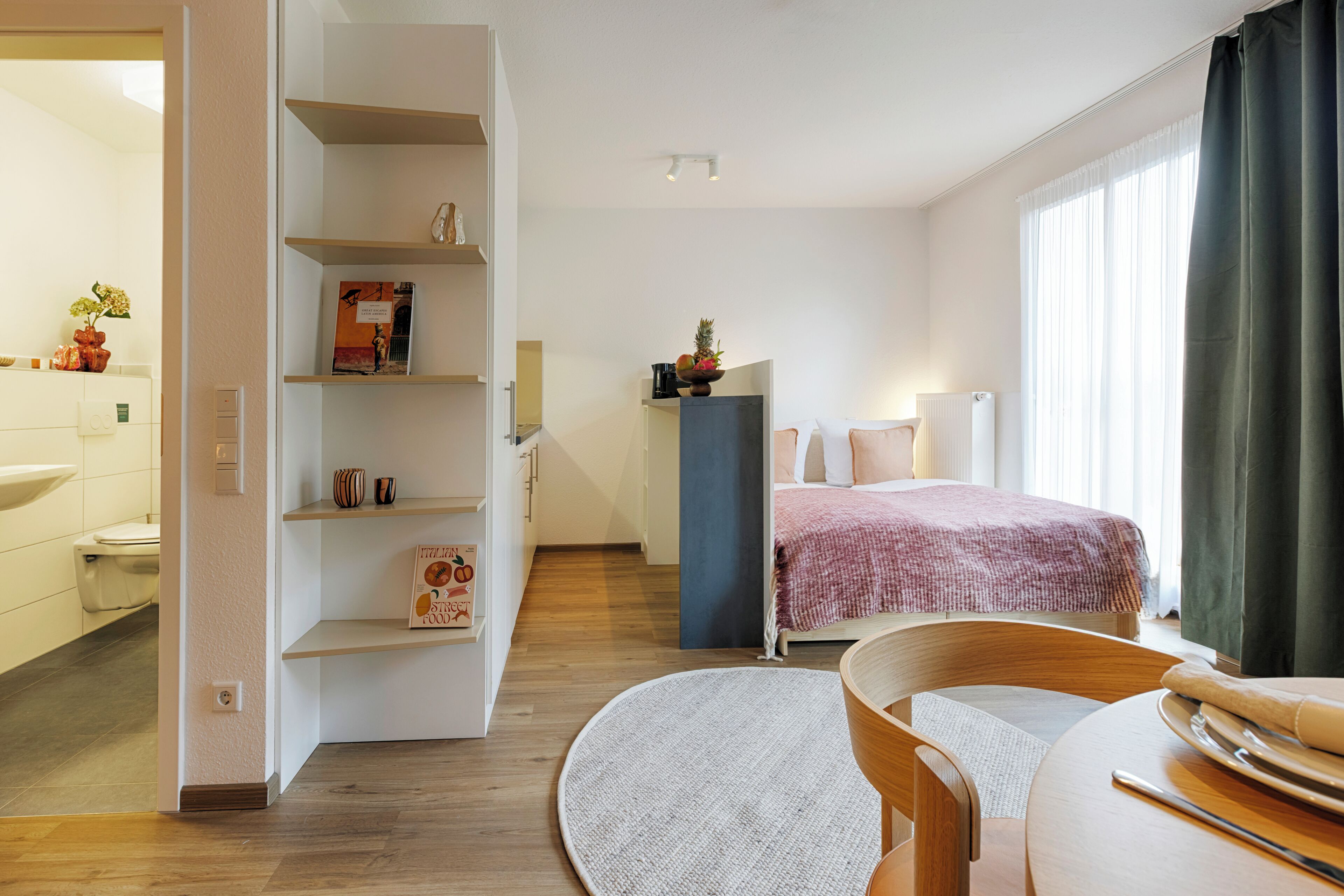 Ein modernes Hotelzimmer mit Bett, rundem Esstisch, integriertem Bücherregal, Heizkörper und einem Fenster mit Vorhängen.