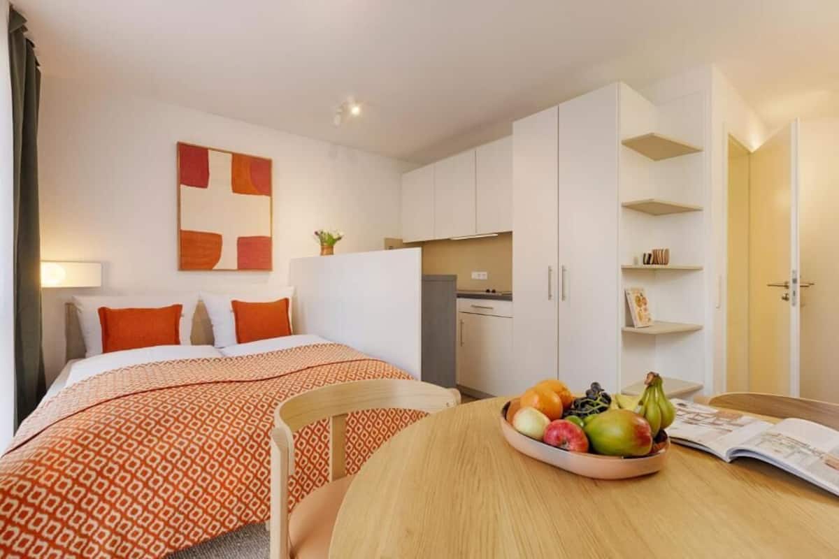 A hotel room with two beds, a dining table with fruit, a kitchen area, and a bookshelf.