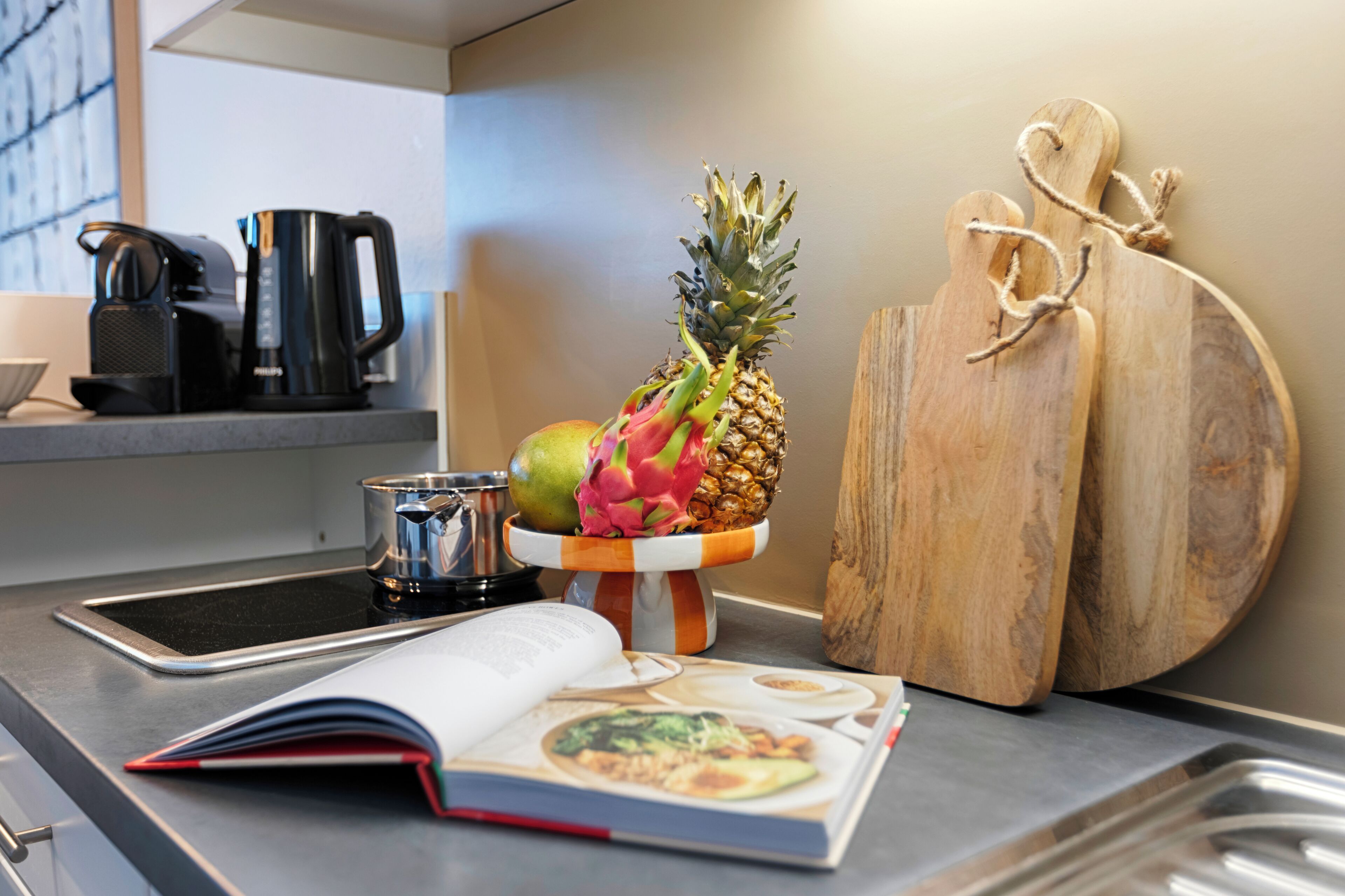 A kitchen counter with a coffee maker, a book, a pineapple, and a cutting board.