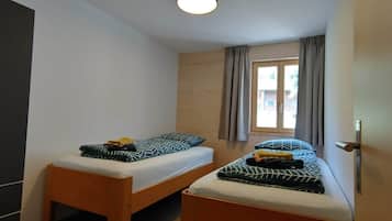Ein kleines, schlichtes Zimmer mit zwei Einzelbetten, einem Fenster mit Vorhängen und einer Tür.