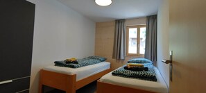 A small, simple room with two single beds, a window with curtains, and a door. - Chalet Mallum – Lower Apartment (Grengiols)