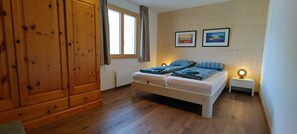 A bedroom with a wooden wardrobe, a bed with patterned bedding, two bedside tables with lamps, and two framed pictures on the wall. - Chalet Mallum – Lower Apartment (Grengiols)