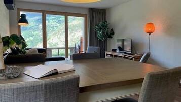 Ein modernes Wohnzimmer mit einem großen Esstisch, bequemen Sitzmöglichkeiten, einem Fernseher und Blick ins Freie.