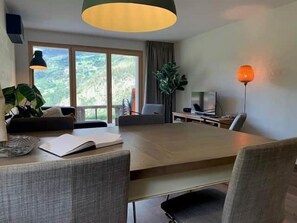 A modern living room with a large dining table, comfortable seating, a television, and a view of the outdoors. - Chalet Mallum – Lower Apartment (Grengiols)