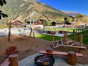 Ein Spielplatz mit Schaukeln und einer Feuerstelle, umgeben von Holzelementen und einer Bergkulisse.