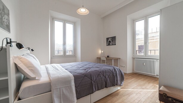 Un dormitorio con cama, escritorio y dos ventanas.