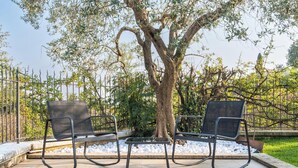 Deux espaces de détente en plein air, avec des chaises et des tables en métal, entourés d’un arbre et d’une clôture en métal.