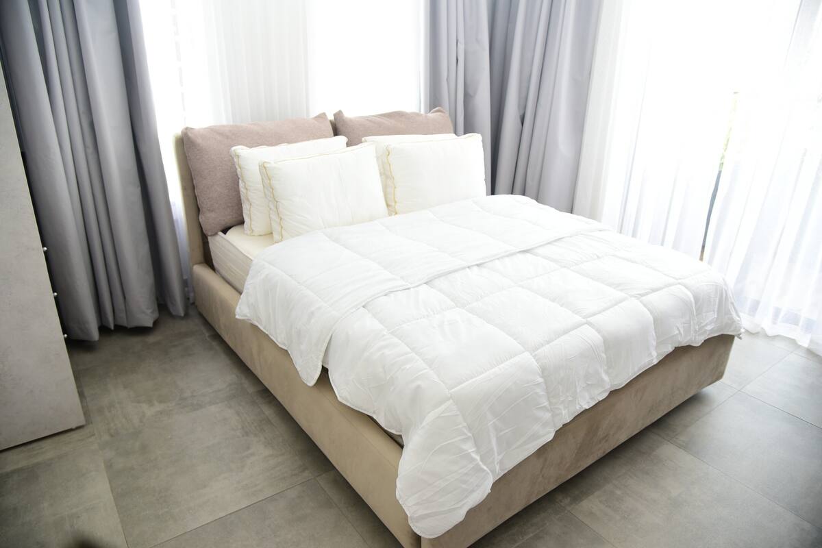 Un lit avec une couette blanche, des oreillers beige et des rideaux gris, dans une pièce dont le sol est recouvert de carreaux.