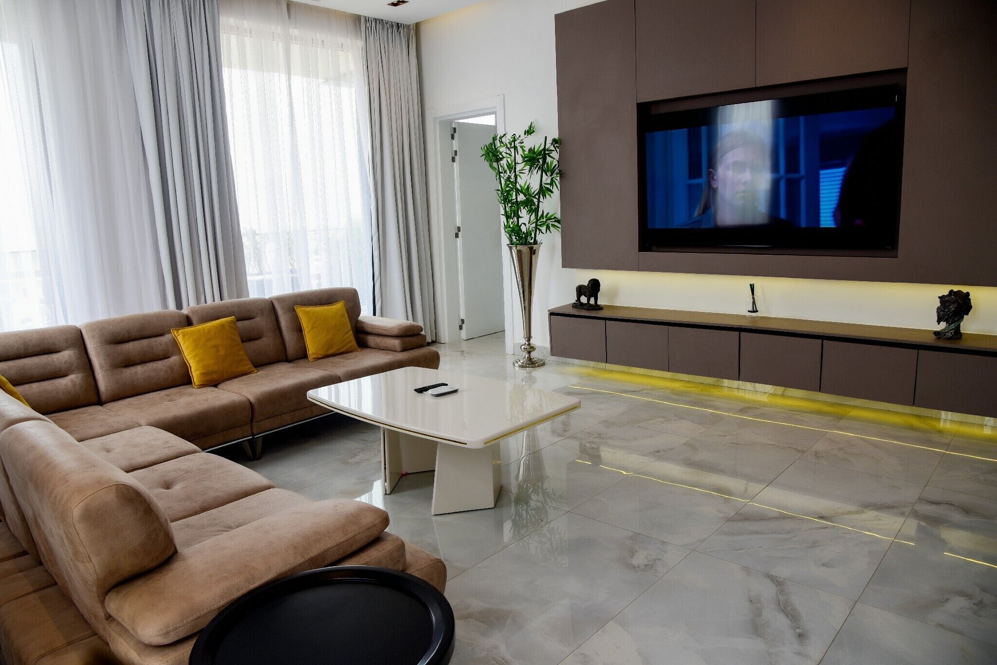 A modern living room with a sectional sofa, a coffee table, a flat-screen TV mounted on the wall, and a potted plant.