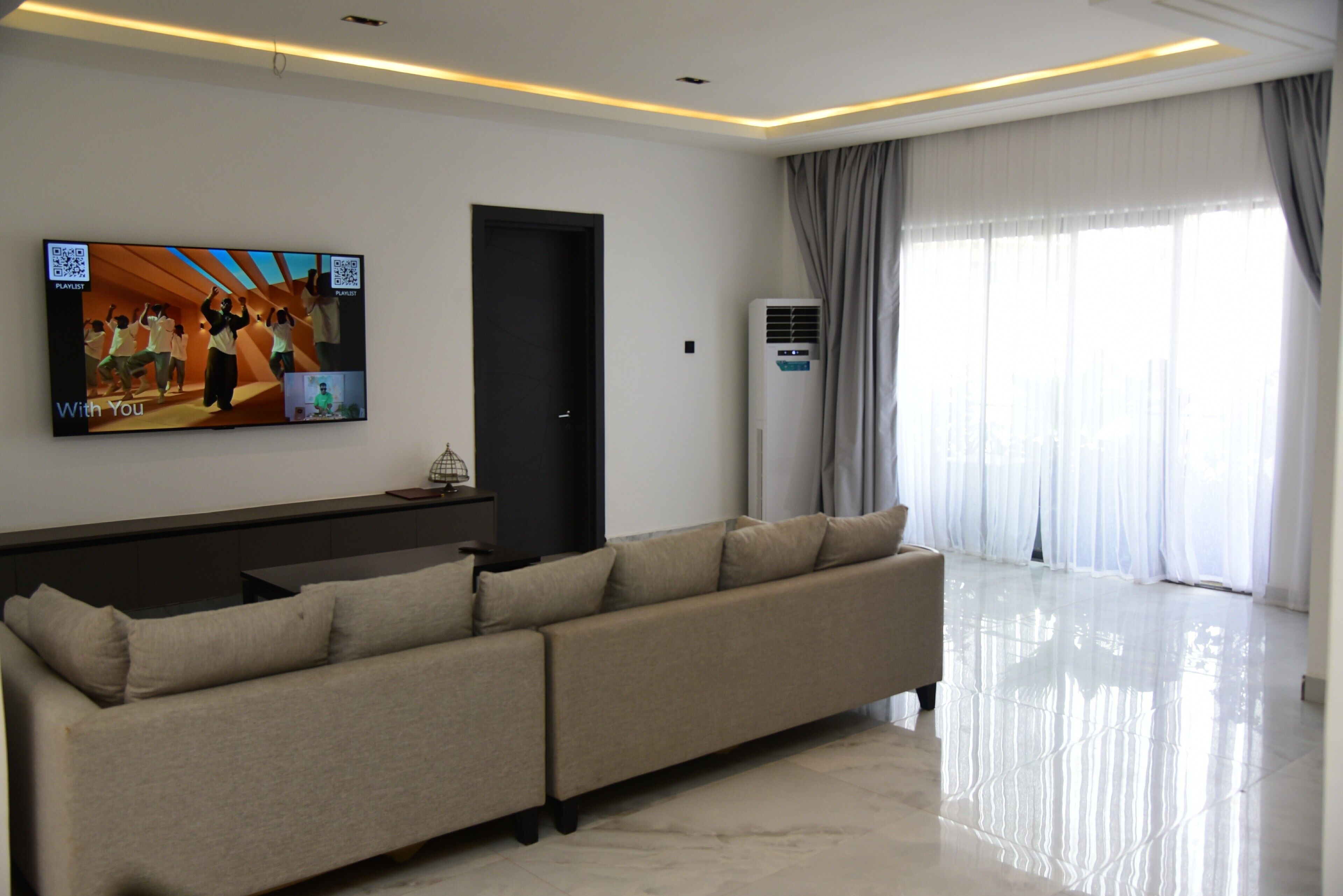 A modern living room with a large flat-screen TV, a sectional sofa, a door, and a window with curtains.