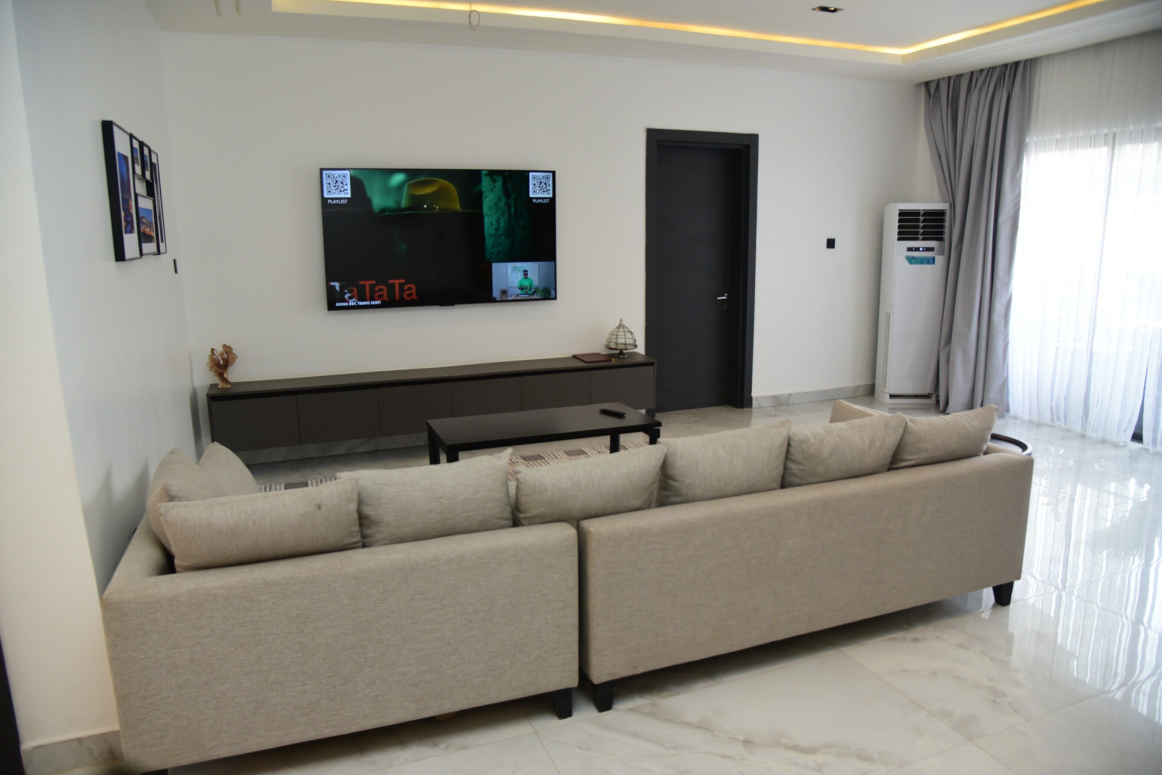 A modern living room with a sectional sofa, a flat-screen TV, a dark-colored console table, and a large air conditioning unit.