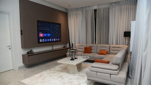 Egy modern nappali, melyben egy nagy, falra szerelt, lapos képernyős televízió, egy szürke kanapé narancssárga párnákkal, egy fehér dohányasztal és egy mintás szőnyeg található.