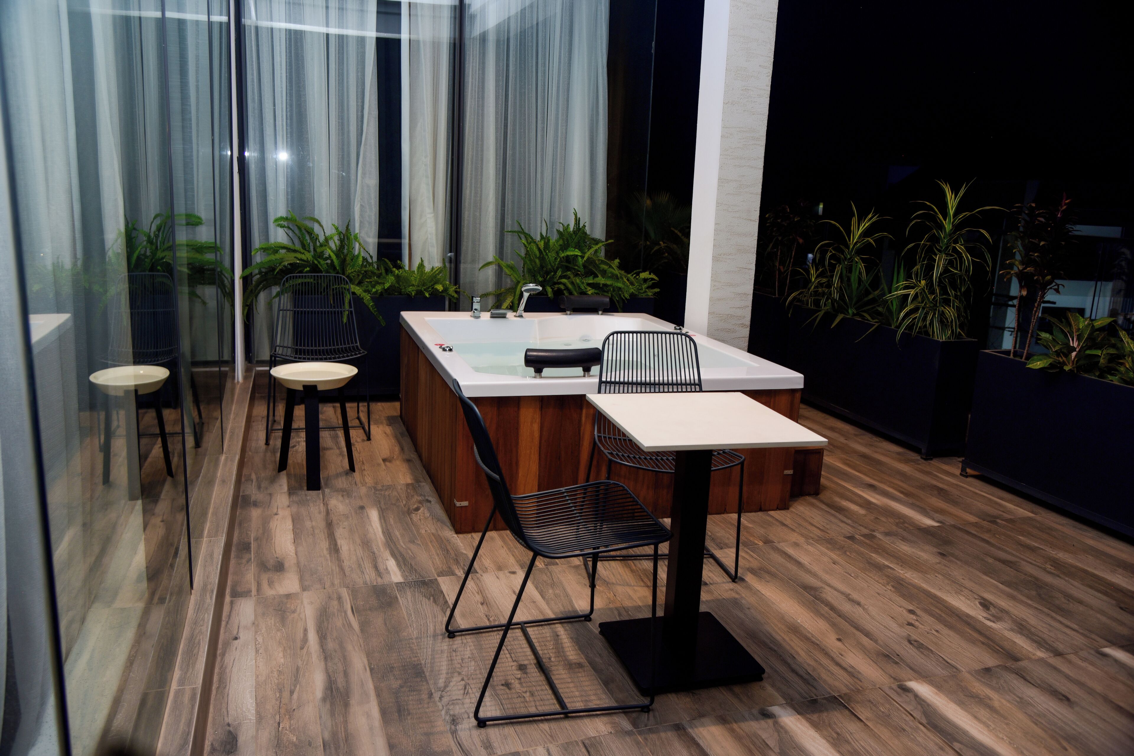 A modern outdoor dining area with a wooden floor, a white table, black chairs, and a kitchen island with a sink.