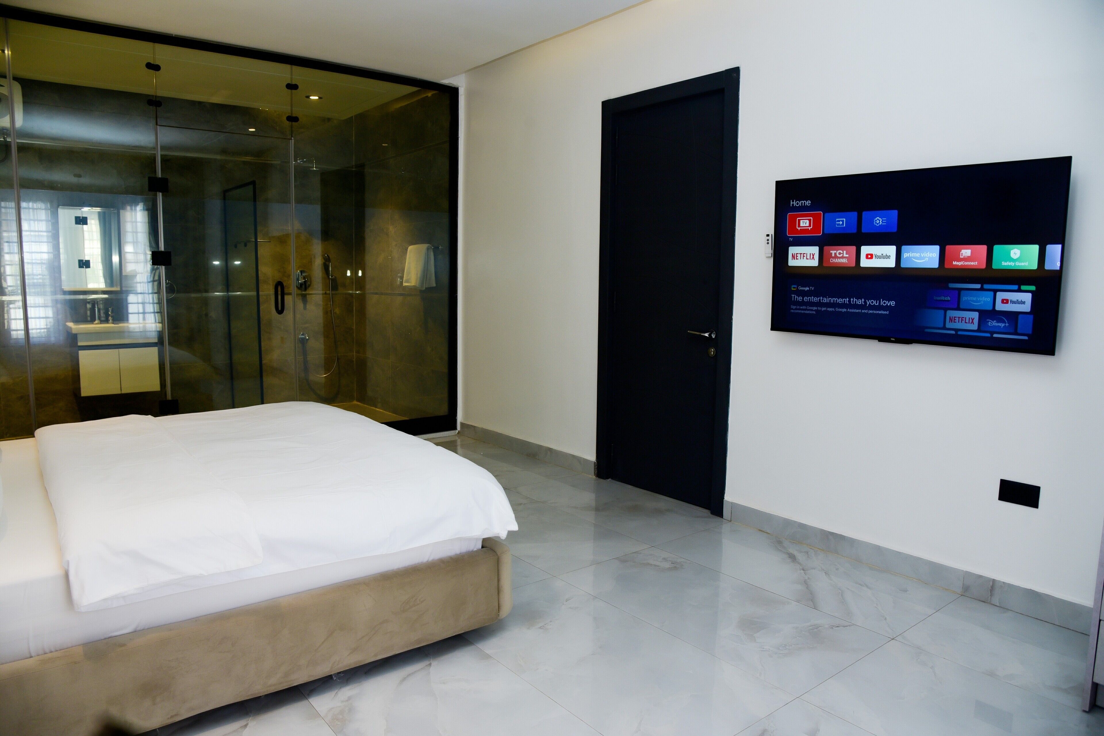 Une chambre d’hôtel moderne équipée d’un lit, d’une douche en verre, d’une porte noire et d’un téléviseur fixé au mur qui permet d’accéder à différents services de diffusion en continu.