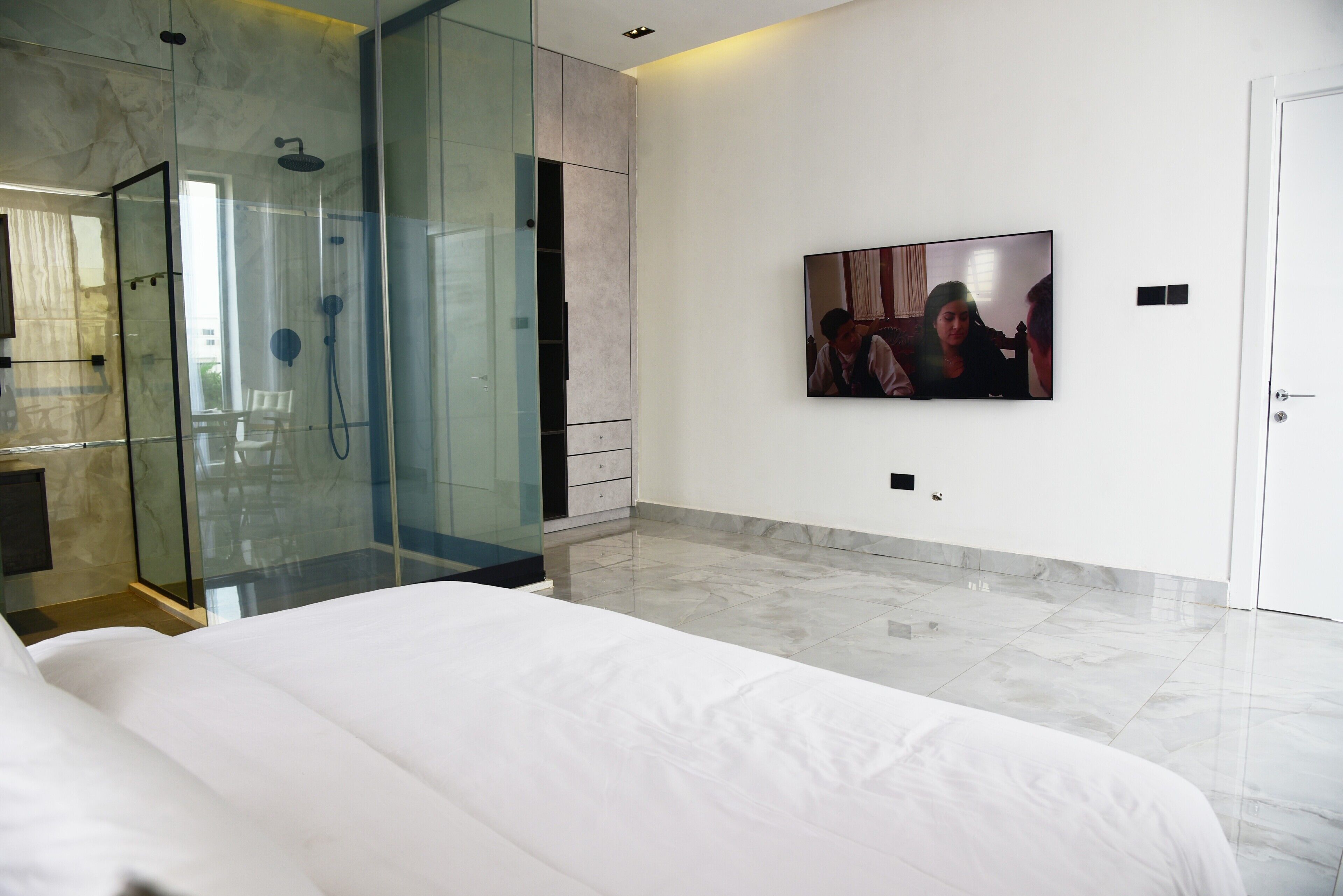 A modern hotel room with a glass-enclosed shower, a large bed, a flat-screen TV mounted on the wall, and a marble floor.