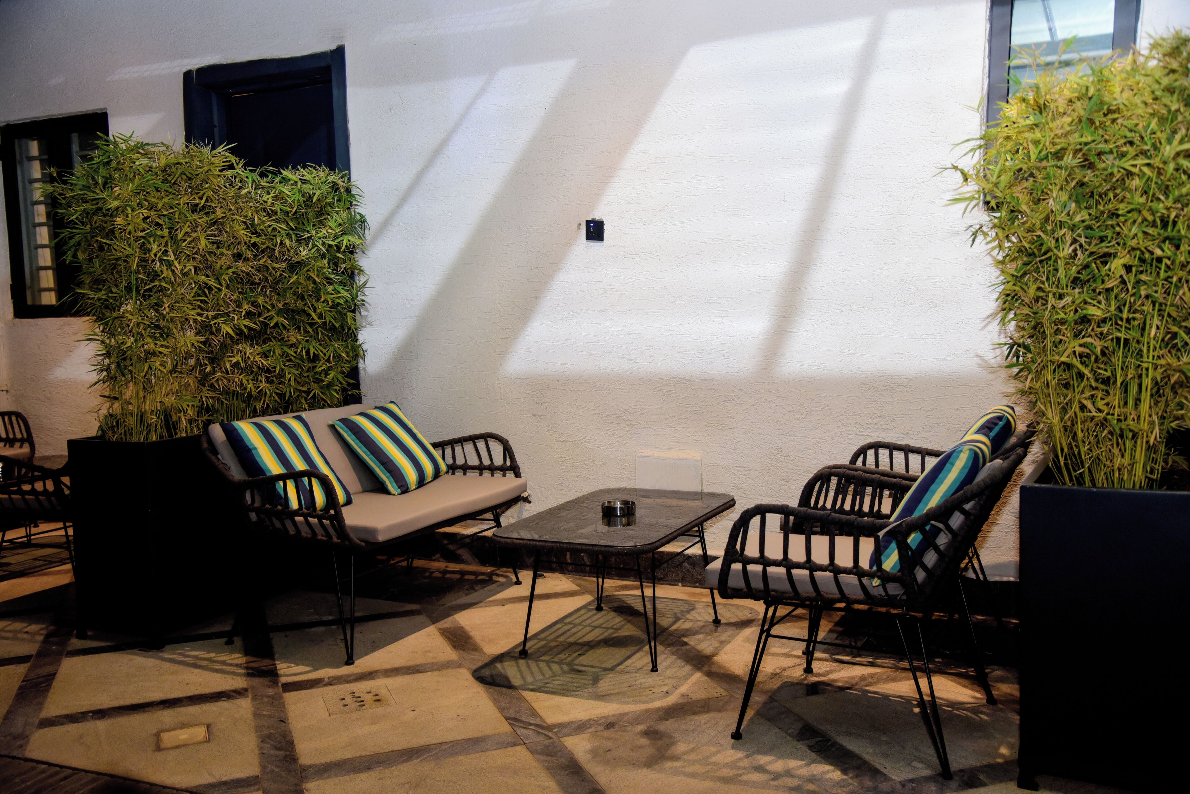 An outdoor seating area with a sofa, chairs, and a table, surrounded by plants and a white wall.
