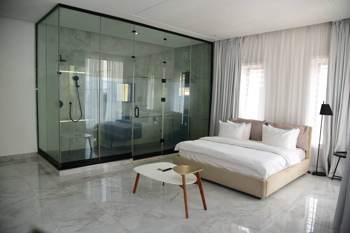 Une chambre moderne avec une douche en verre, un lit avec une literie blanche et une petite table blanche.