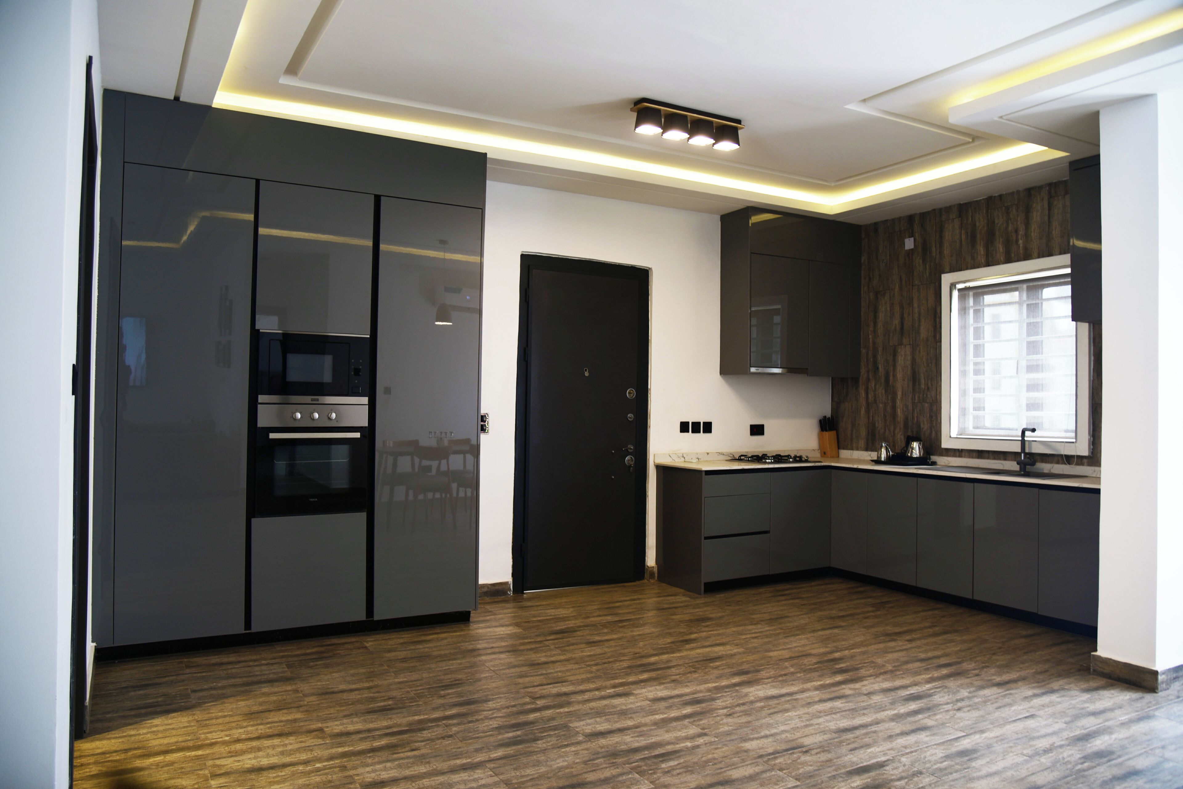 Une cuisine moderne avec des meubles de couleur sombre, un four encastré et une fenêtre équipée de stores.