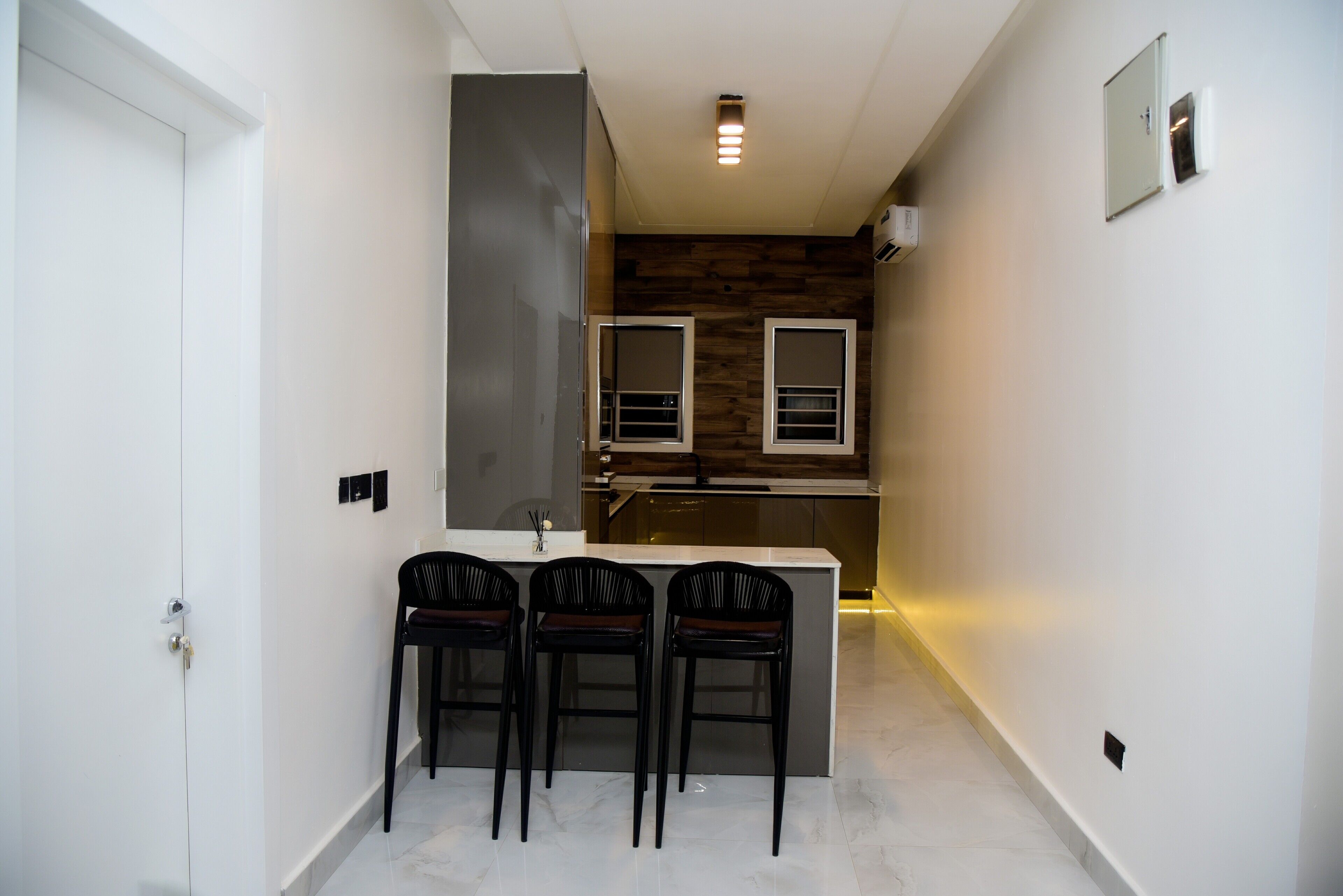 A narrow hallway leading to a kitchen with a wooden accent wall, a white door, and three black bar stools.