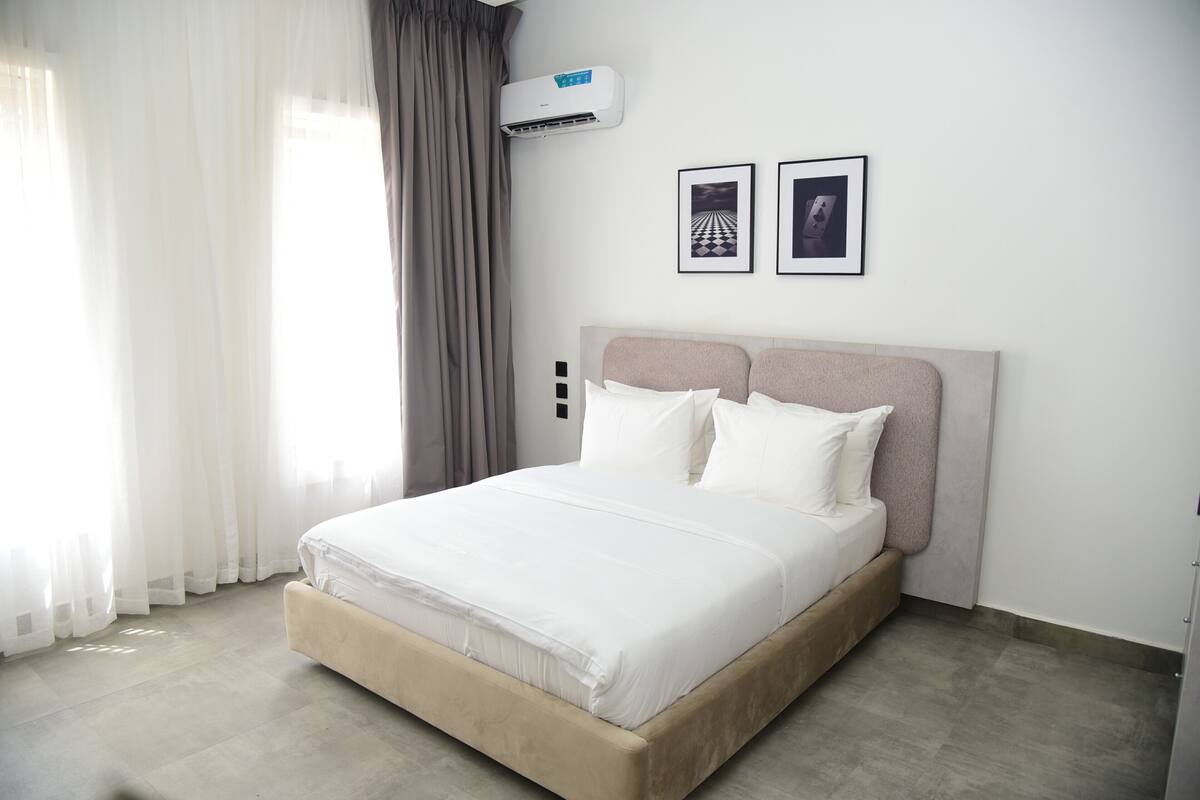 Un lit bien fait dans une chambre d’hôtel, avec une tête de lit, un appareil de climatisation et deux tableaux encadrés accrochés au mur.