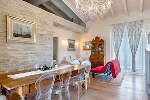 Comedor con mesa de madera y sillas transparentes, una araña y pared de ladrillo.