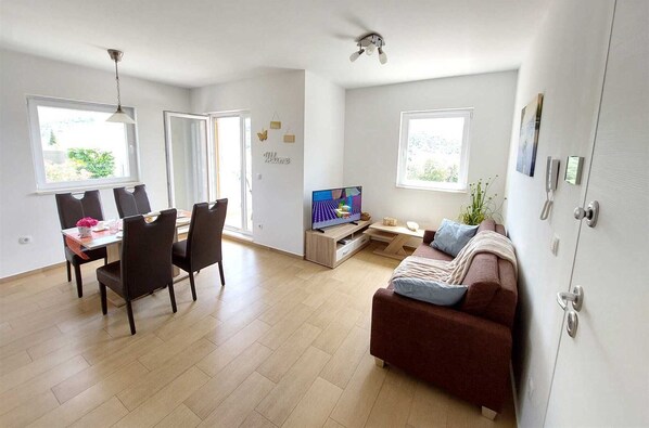 A modern living room with a brown sofa, a dining area with a table and chairs, and a TV on a wooden stand.