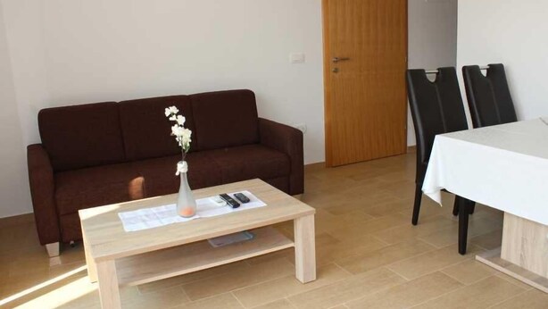 A room with a brown sofa, a wooden coffee table with a vase and remote controls, a white table with chairs, and a wooden door. - A1 - Chic One-Bedroom Apartment with Parking and Garden view (Slano)