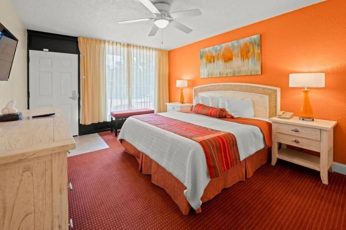 A hotel room with a bed, bedside tables, a ceiling fan, a window with curtains, and a painting on the wall.
