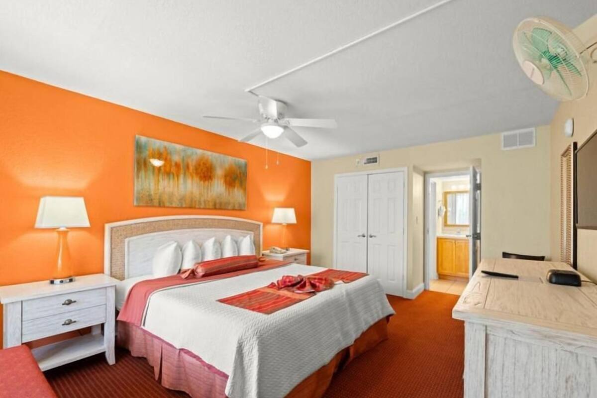 A hotel room with a bed, bedside tables, a ceiling fan, a painting, a door, and a desk with a mirror.